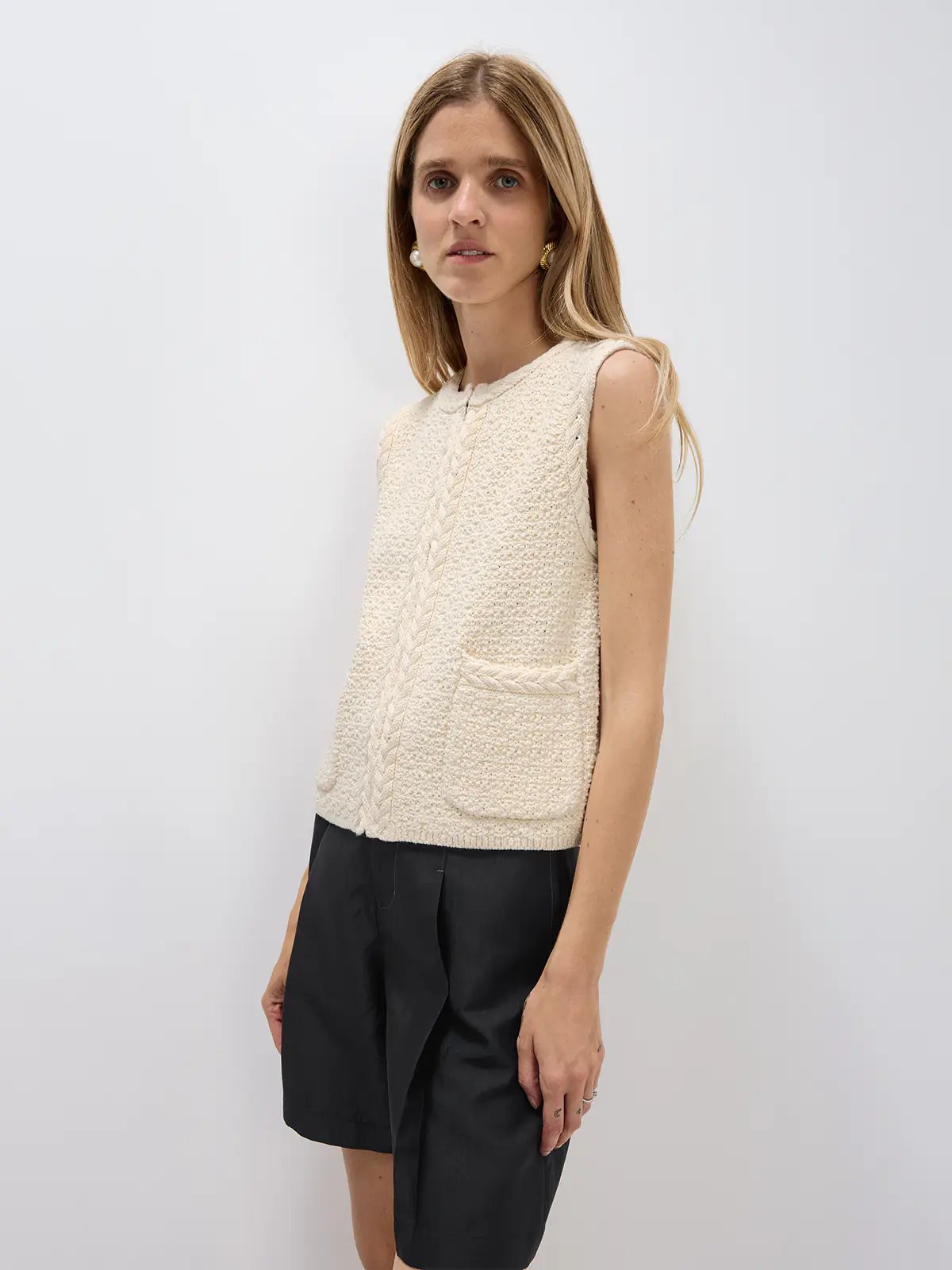 Off-white sleeveless crochet vest with cable trim and patch pockets, shown from the side on a model wearing shorts.