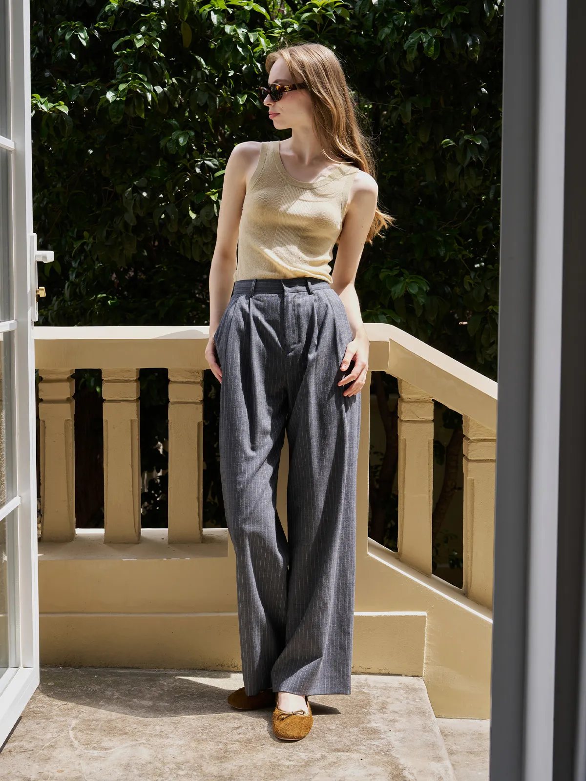 Model in ORAN grey stripe wide leg trousers with beige knit tank and brown ballet flats on outdoor balcony
