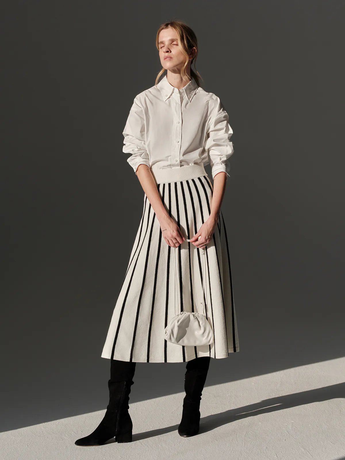 Side view of a model in a Black/White vertical stripe A-line knit skirt with white waistband, white shirt, and black boots.