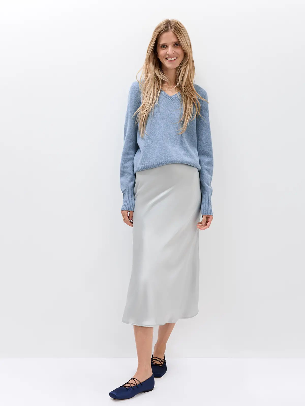 Front view of a model in a blue Merino Wool V-Neck Jumper with a satin midi skirt and lace-up flats.