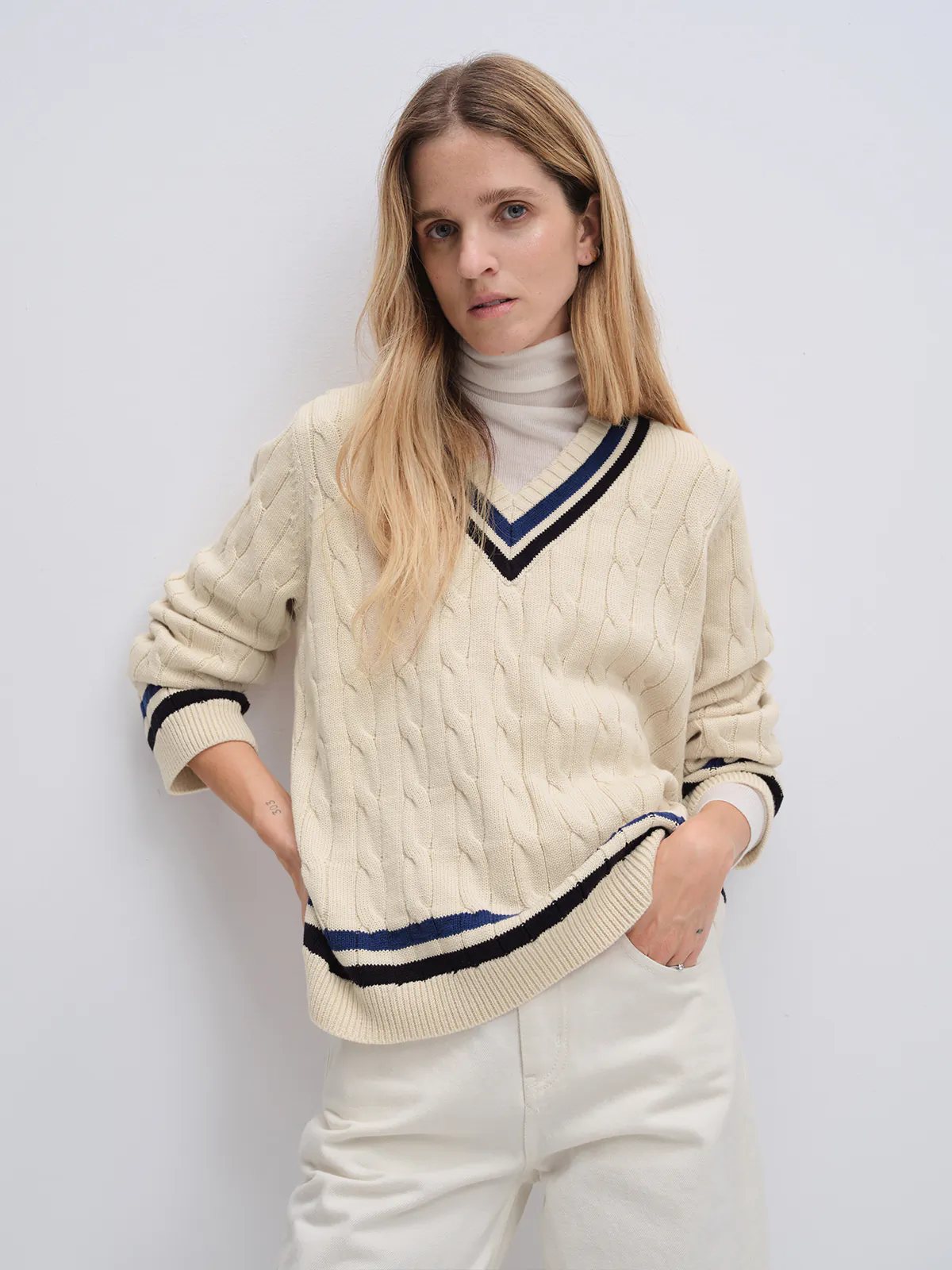 Off-white cable knit V-neck tennis jumper with contrasting trim, paired with wide-leg trousers and loafers, hands on hips.