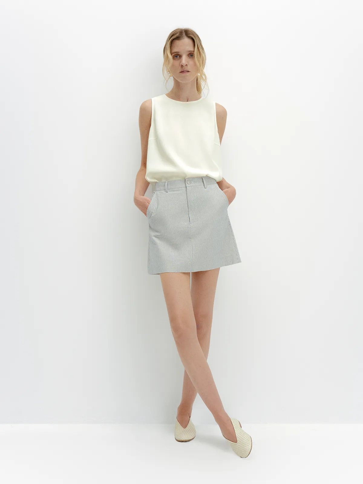 Blonde model wearing ivory MADGE blouse with light gray striped mini skirt and woven flats, hands in pockets