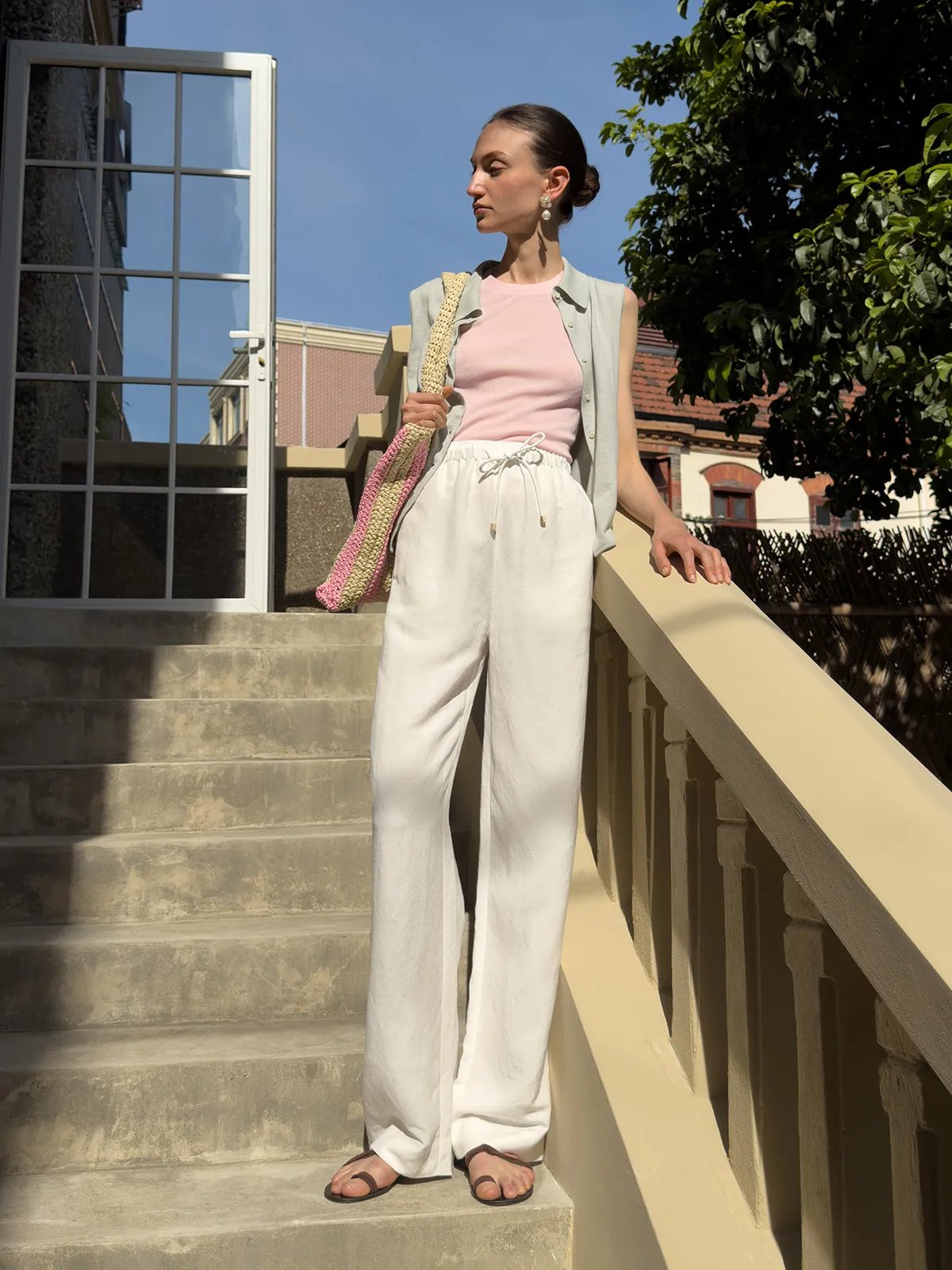 Model wearing ivory linen wide-leg trousers with pink tank, grey vest, and striped tote on outdoor stairs