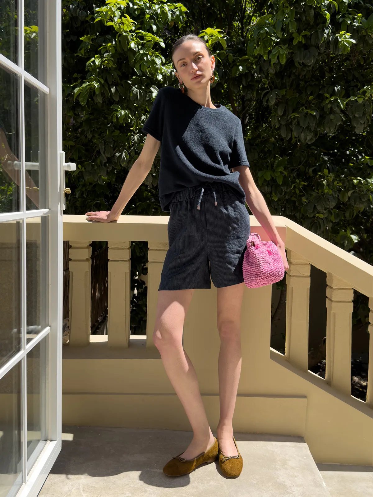 Model posing on sunlit balcony in NEVAEH shorts, black tee, pink bag, and mustard flats
