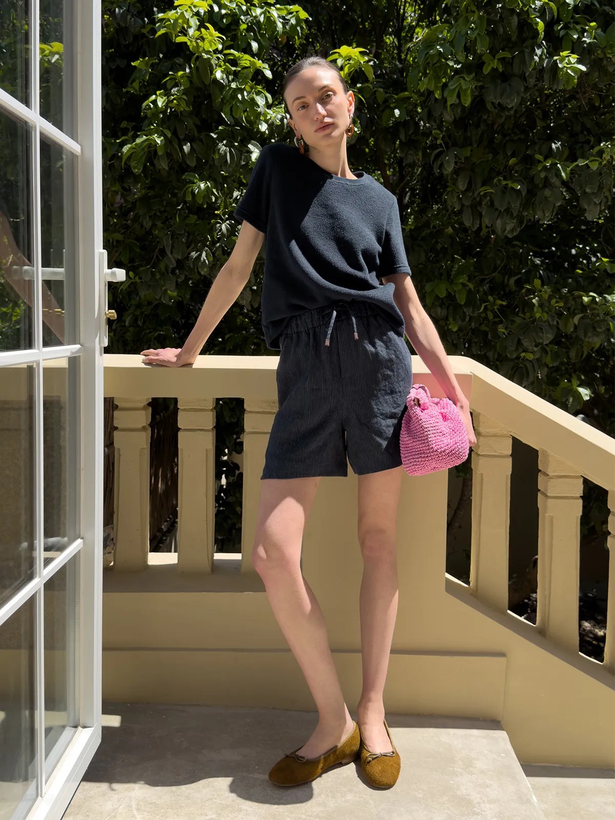 Model posing on sunlit balcony in NEVAEH shorts, black tee, pink bag, and mustard flats