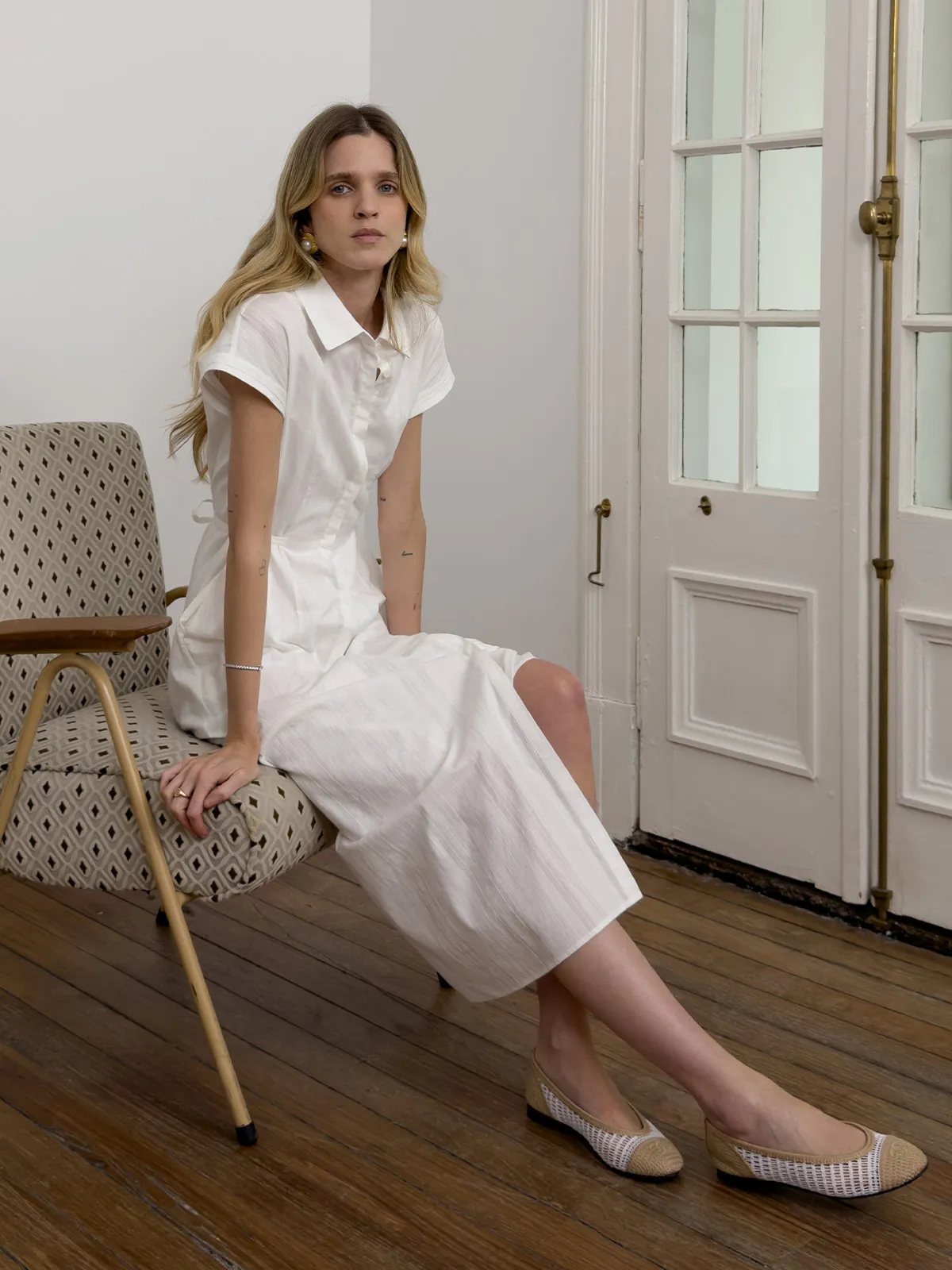 Seated model in white shirt dress with one leg crossed, showing side slit and footwear