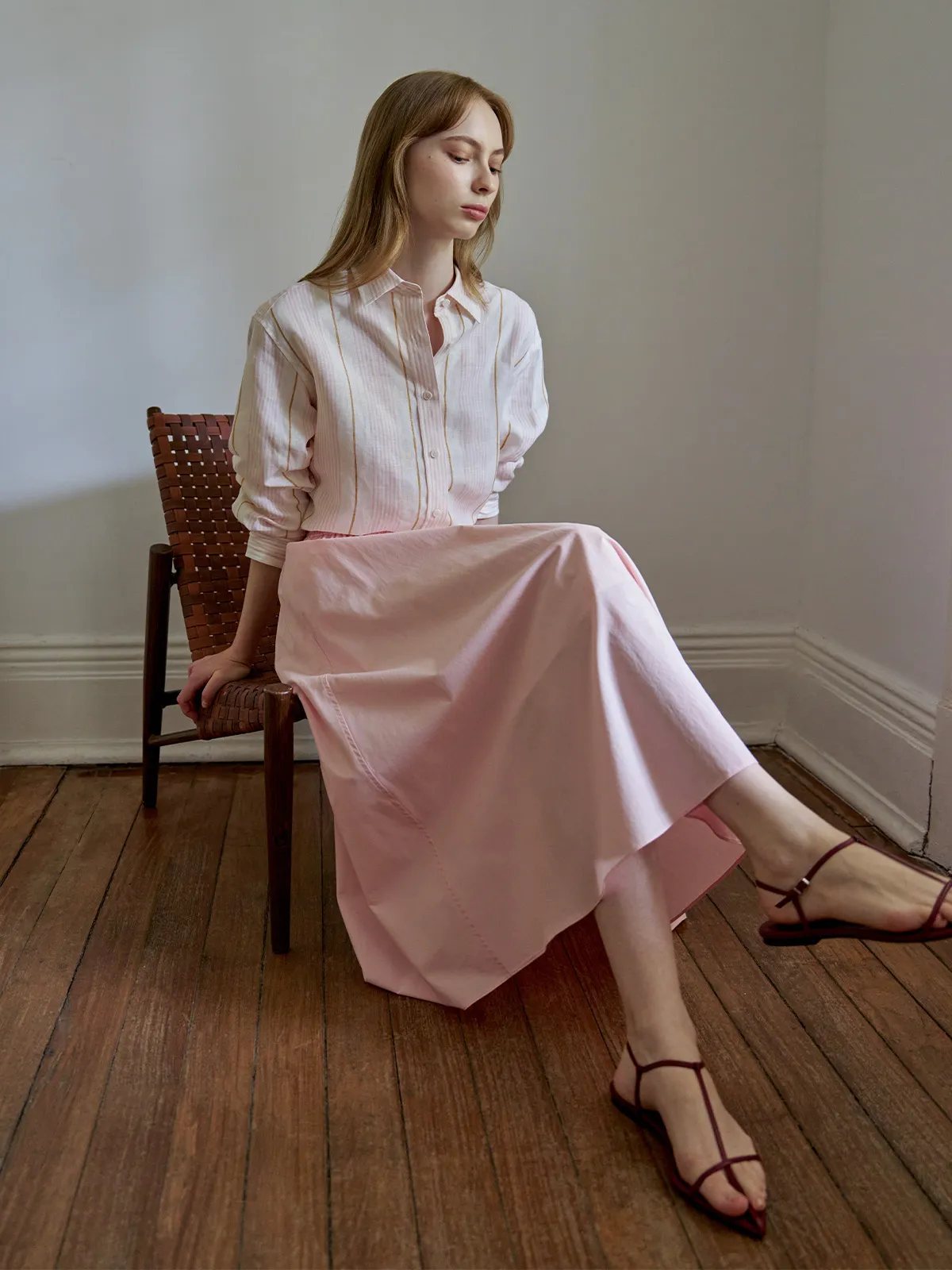 Model seated on woven chair wearing MYRA shirt and pink skirt, indoor minimalist setting
