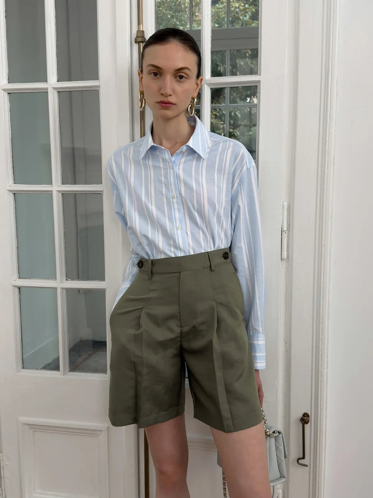 Model wearing blue stripe relaxed shirt tucked into olive green pleated tailored shorts