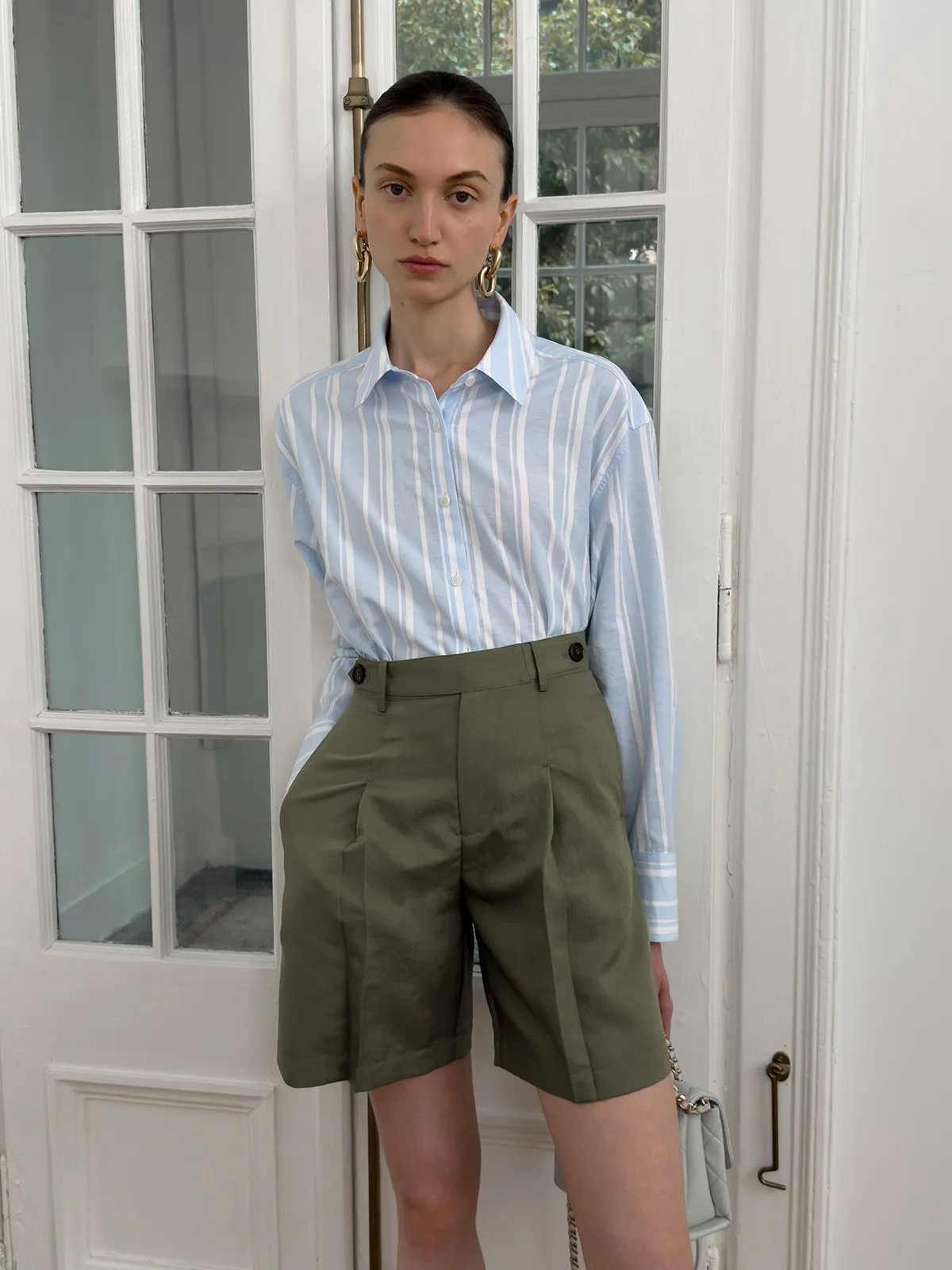 Model wearing blue stripe relaxed shirt tucked into olive green pleated tailored shorts