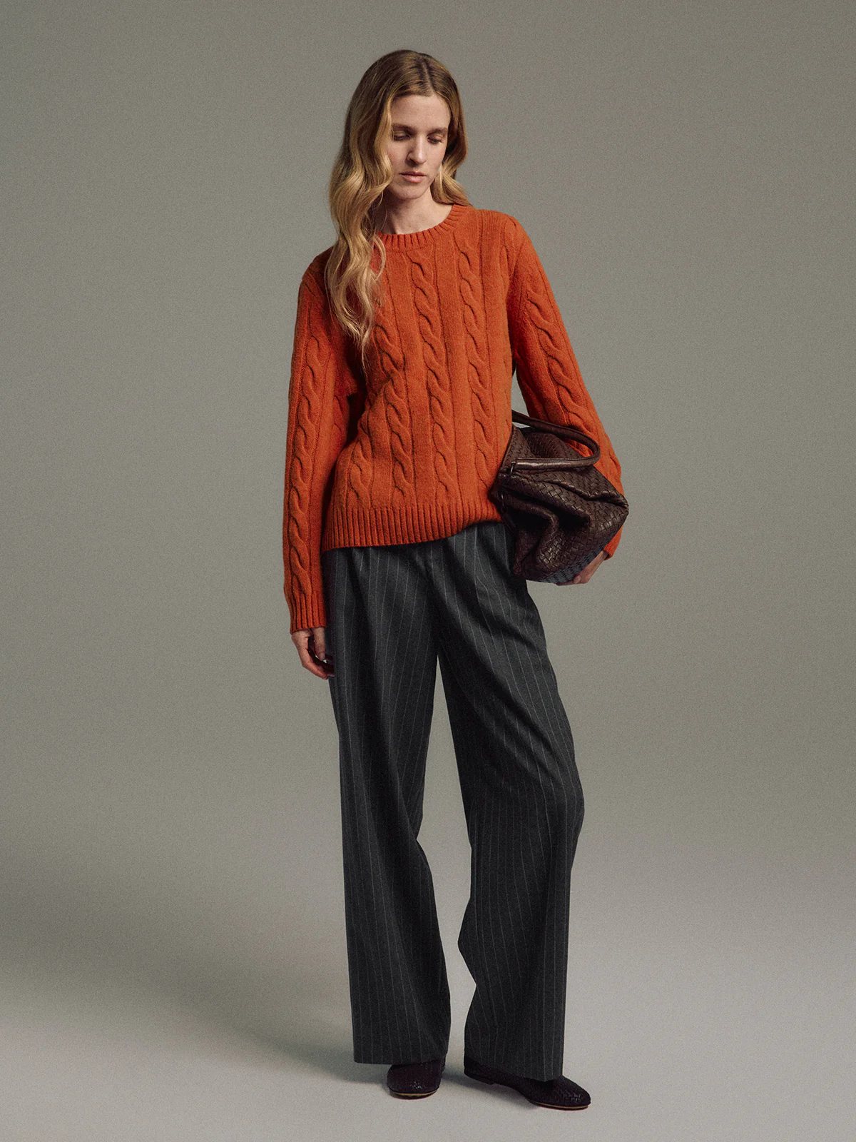 Model wearing an orange cable knit jumper with ribbed hem and cuffs, paired with pinstripe trousers and holding a woven clutch.