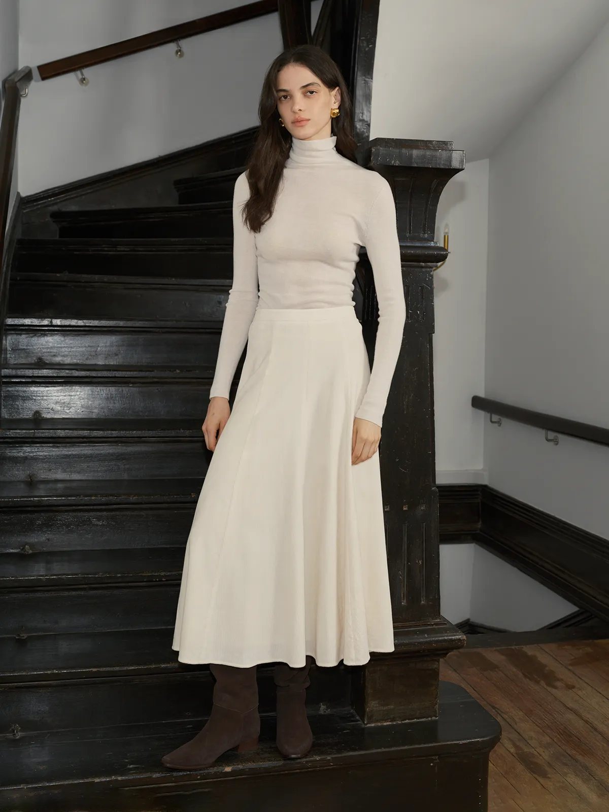 Model standing on dark wooden staircase wearing MILIAN maxi skirt with white turtleneck and brown boots