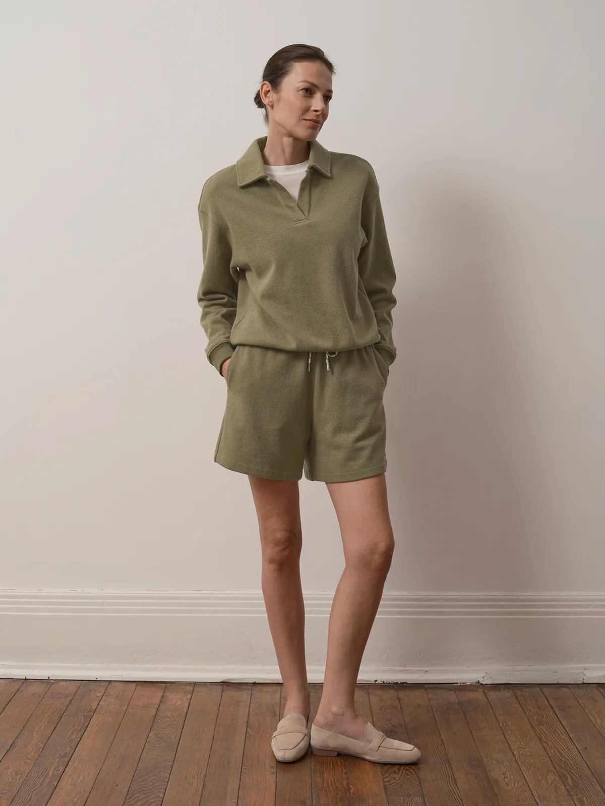 Model in sage green ARLO sweatpants and matching polo-style top, hands in pockets, against white wall and wooden floor