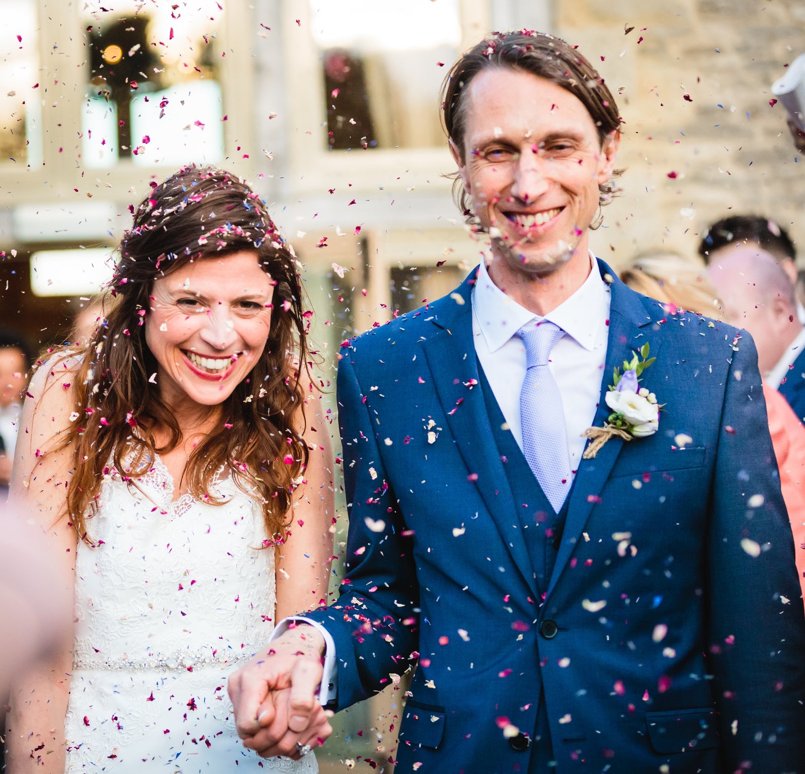 Bride and groom laughing as pink and white confetti falls around them, the groom in a bright blue suit.