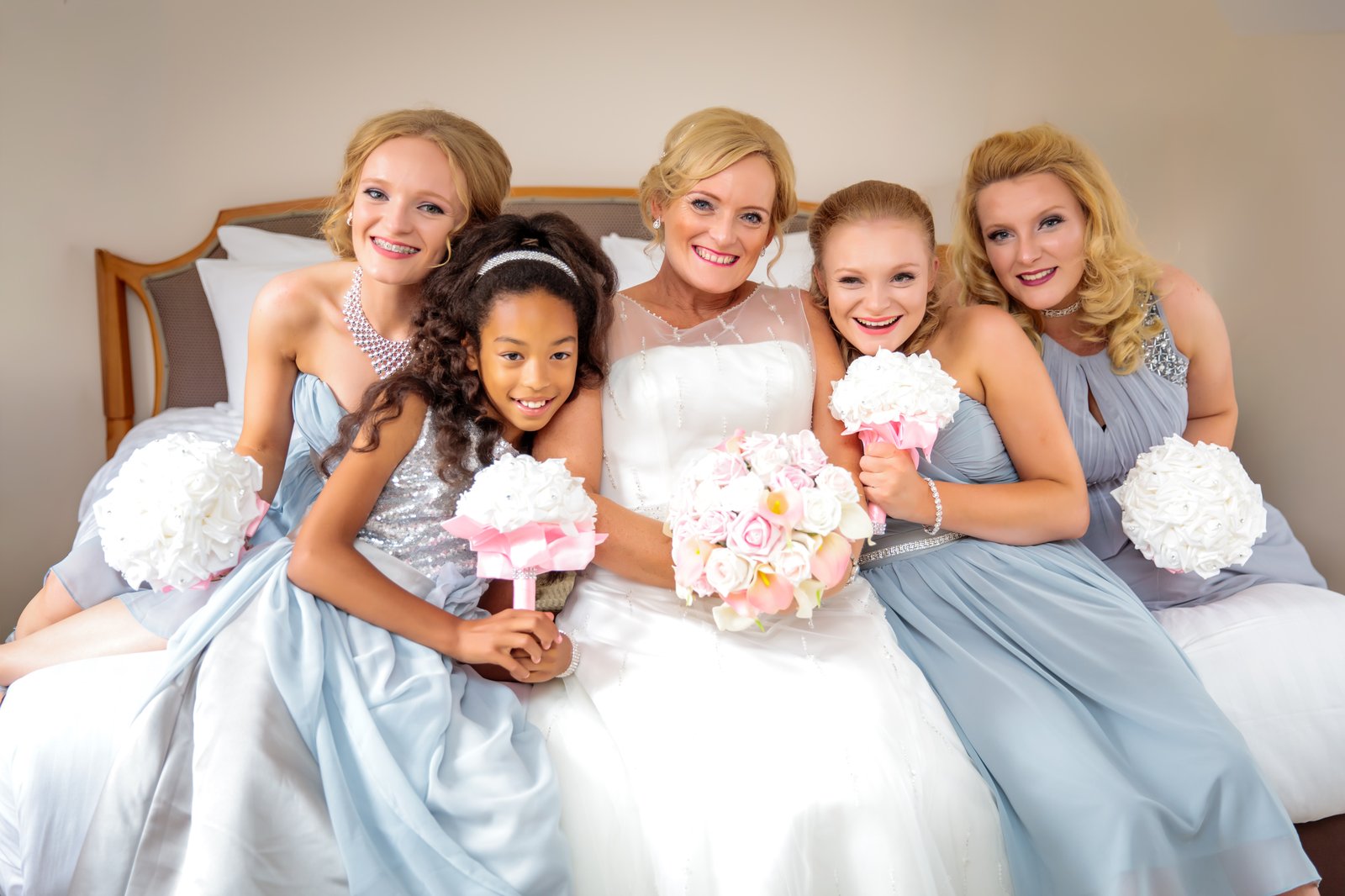 Bride sitting on a bed with her four bridesmaids in pale blue gowns, all holding pastel bouquets.