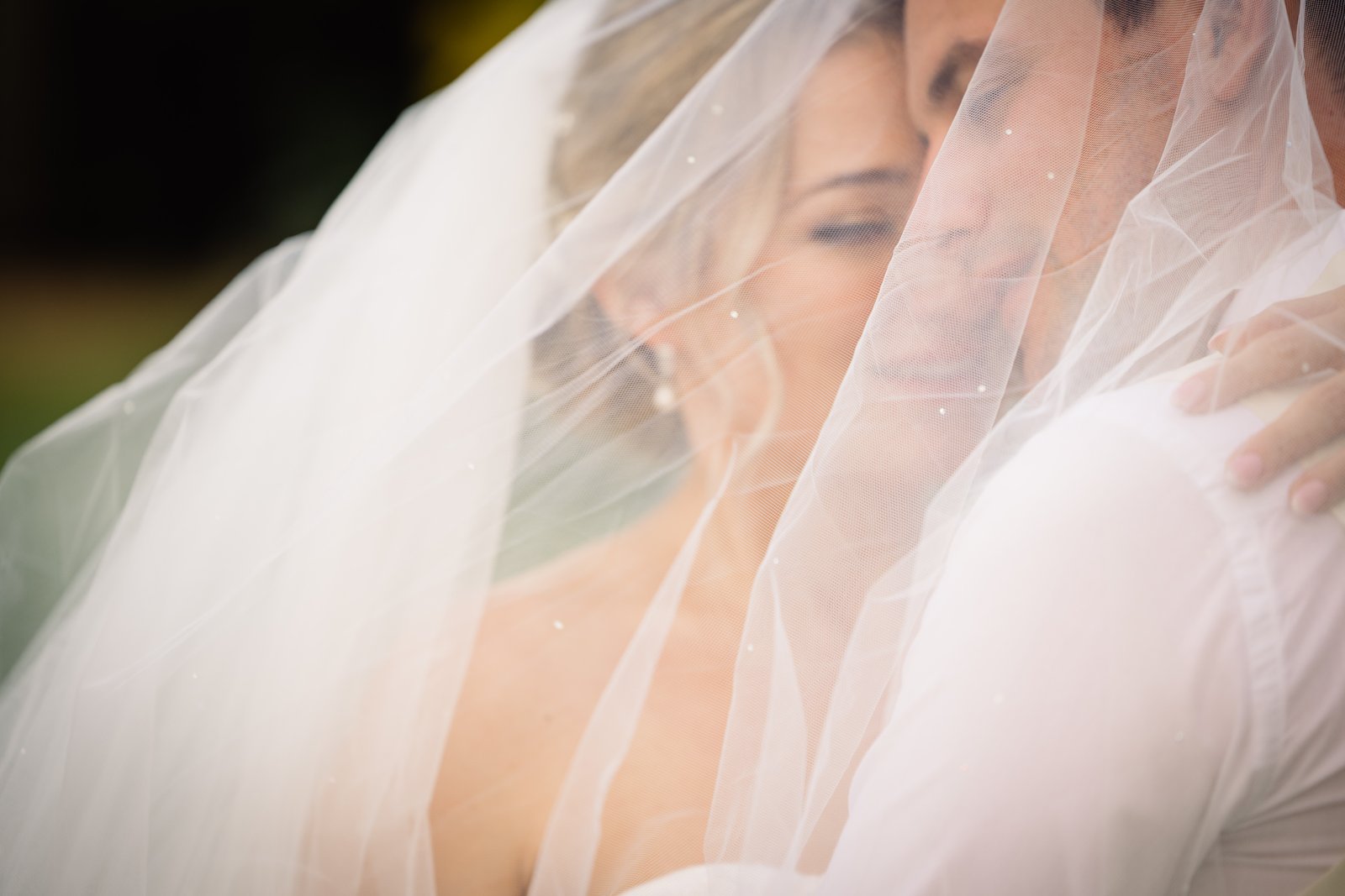 Bride and groom kissing beneath her veil, backlit so the tulle glows around their faces.