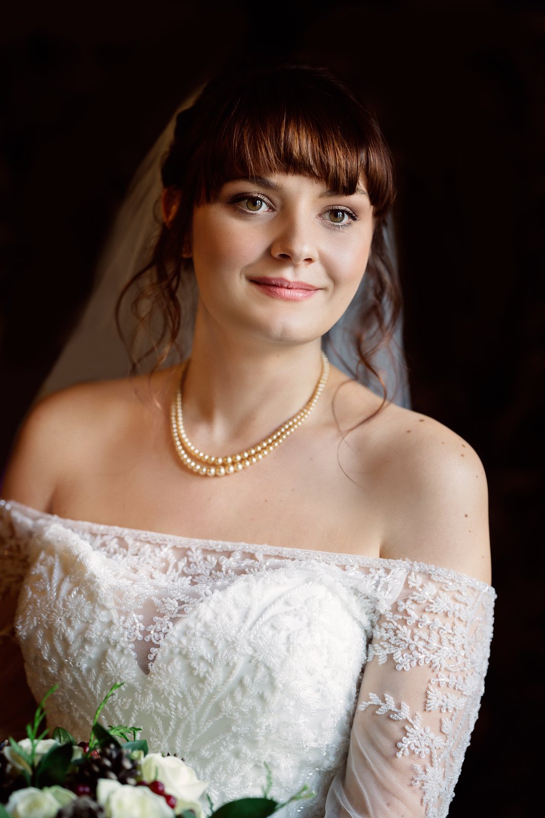 Bride in an off-the-shoulder lace gown and pearl necklace, lit softly against a dark background.