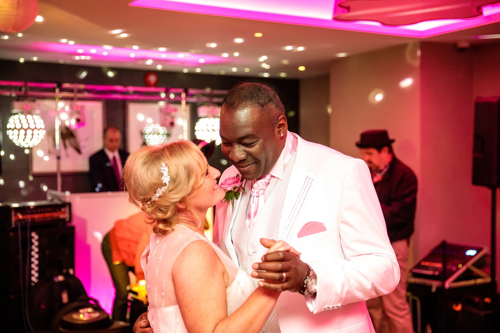 Couple dancing close under pink and magenta uplighting, the DJ booth and disco balls behind them.