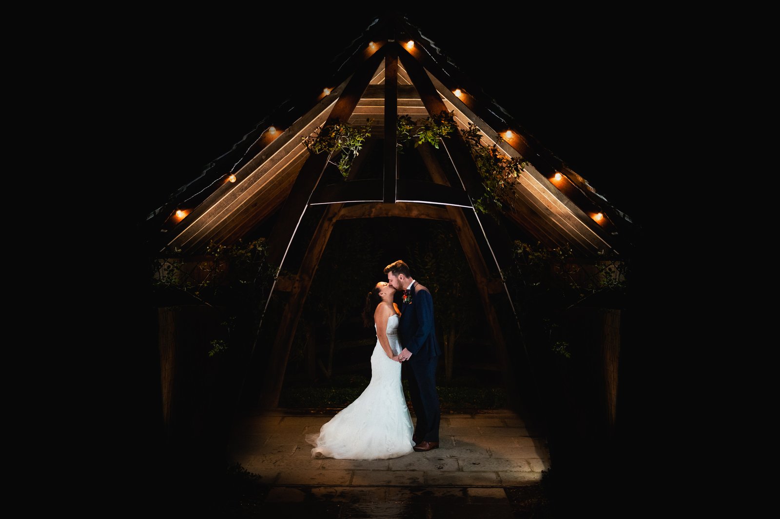 Couple embracing under the lit oak porch at Hyde House after dark, festoon lights strung overhead.