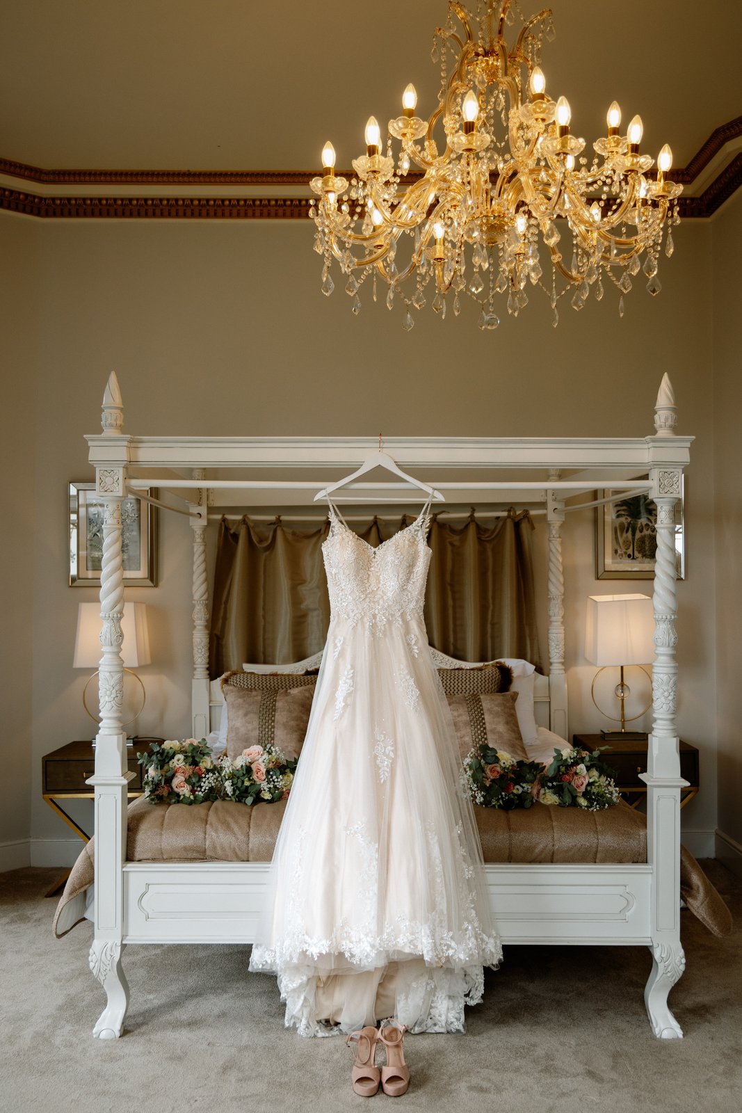 Wedding dress hanging on a white four-poster bed beneath a chandelier in a bridal suite at Bourton Hall.