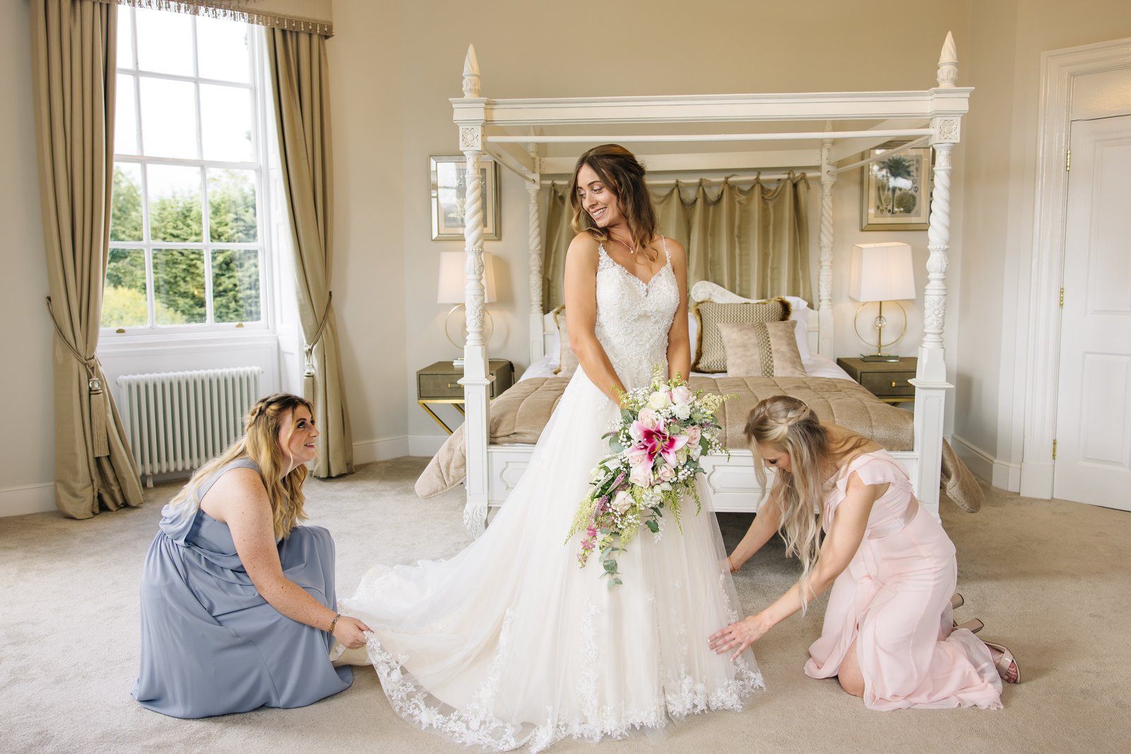 Two bridesmaids kneeling to arrange the bride's train in front of a four-poster bed.