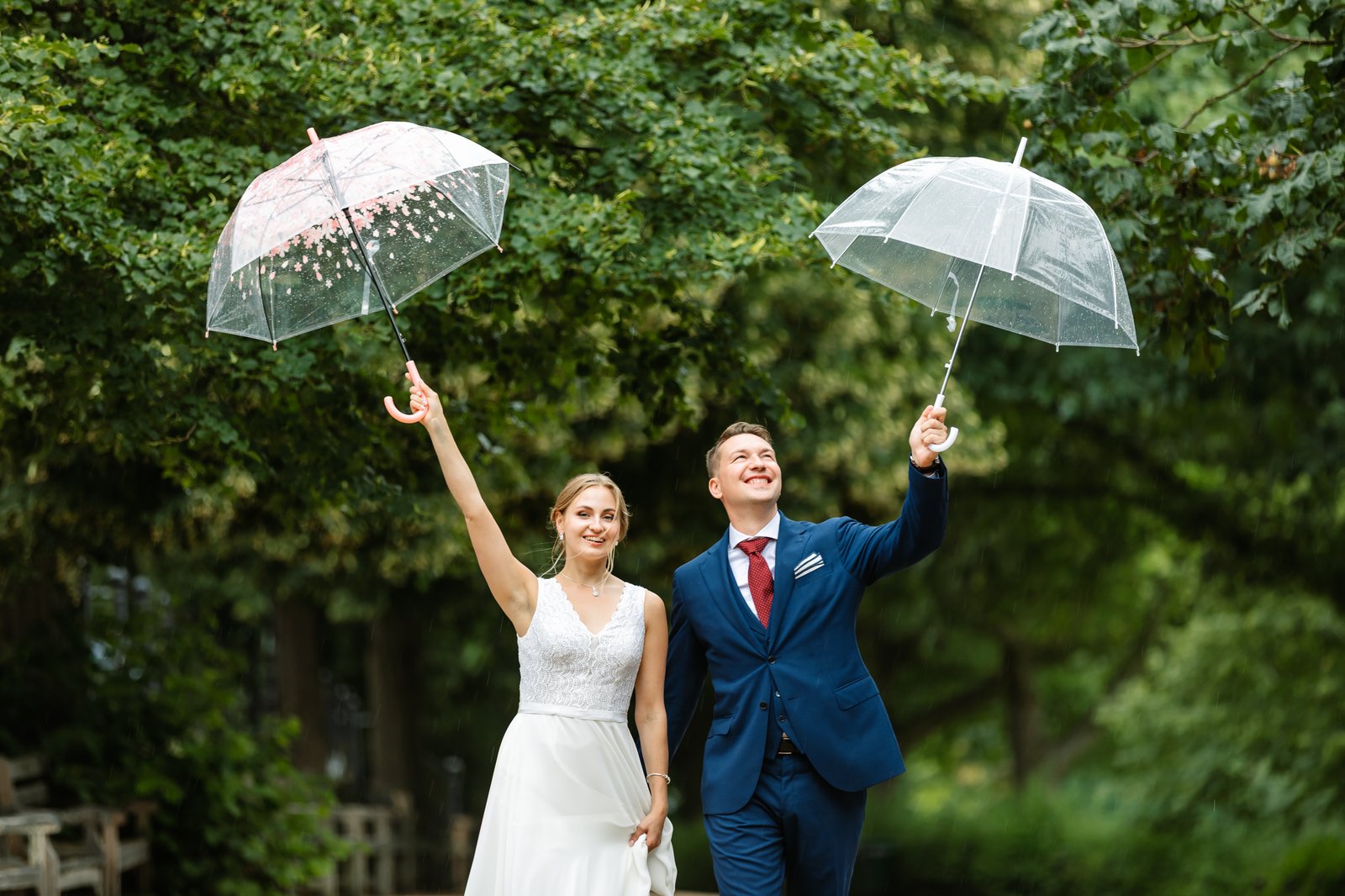 Bride and groom raising clear umbrellas in the rain, laughing under the trees.