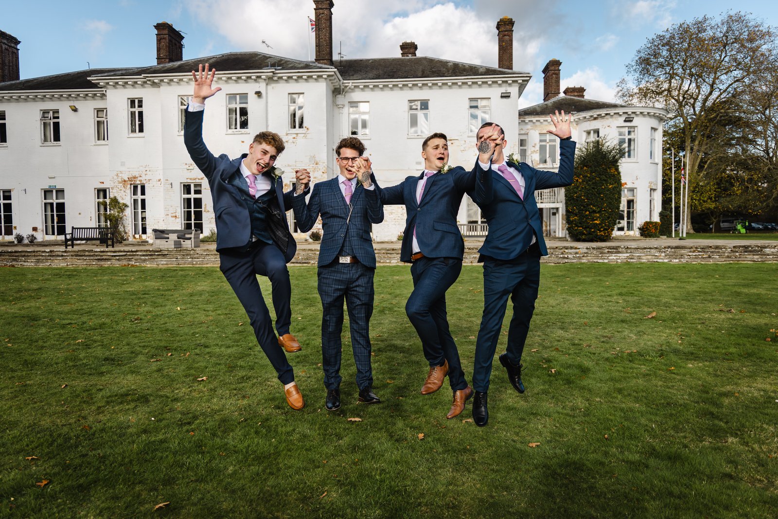 Groomsmen leaping in the air on the lawn in front of a white country house.