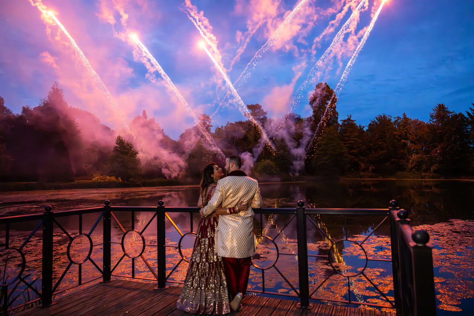 Couple embracing on a deck at The Orangery, Maidstone, as fireworks burst over the lake behind them.