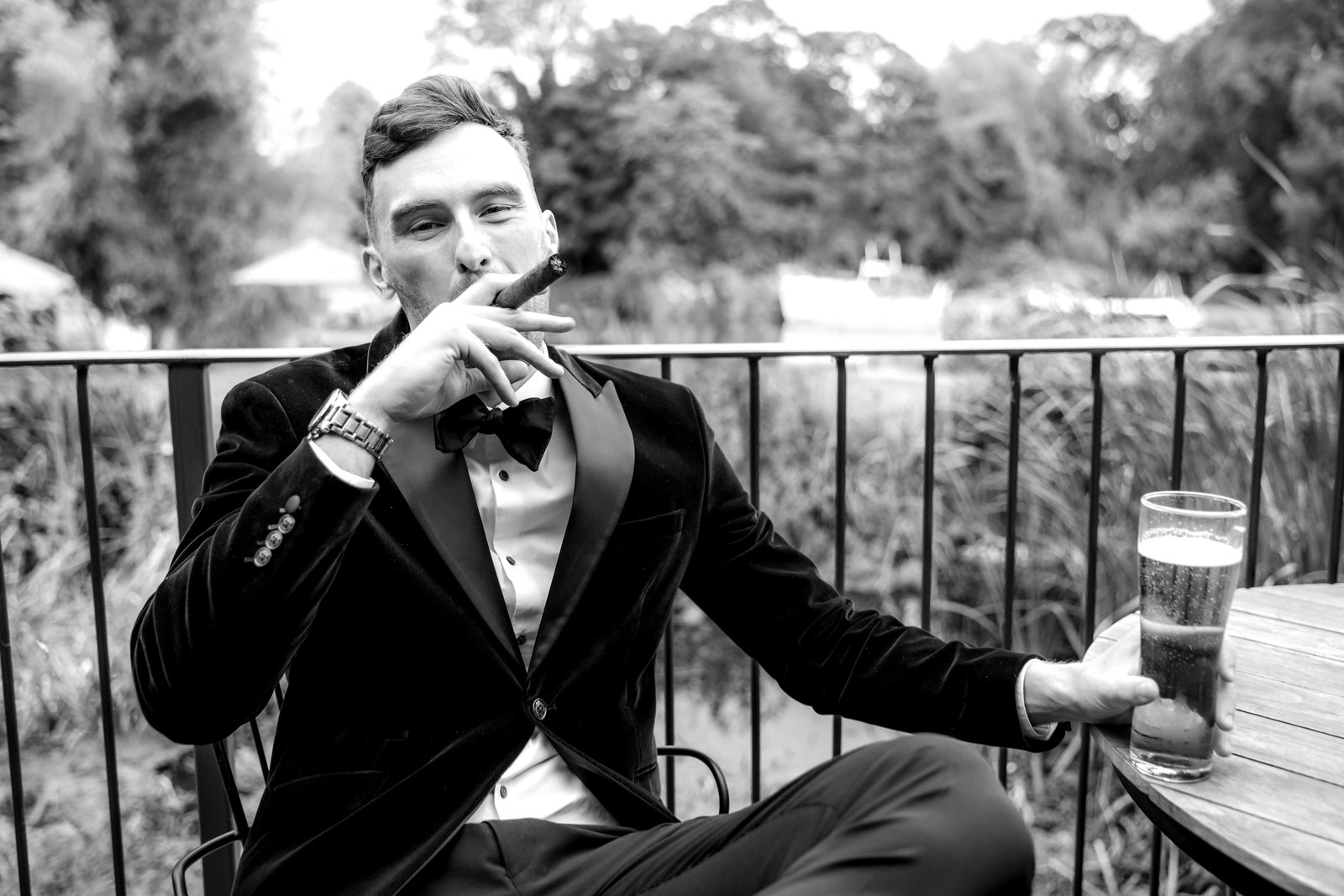 Groom relaxing with a cigar and a pint on the terrace by the river, in black and white.