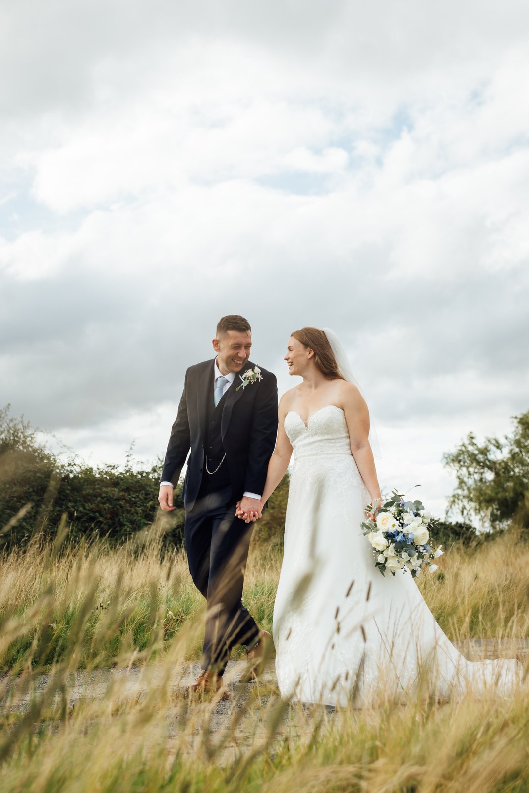 Bride and groom walking hand in hand through long meadow grass under a wide cloudy sky.