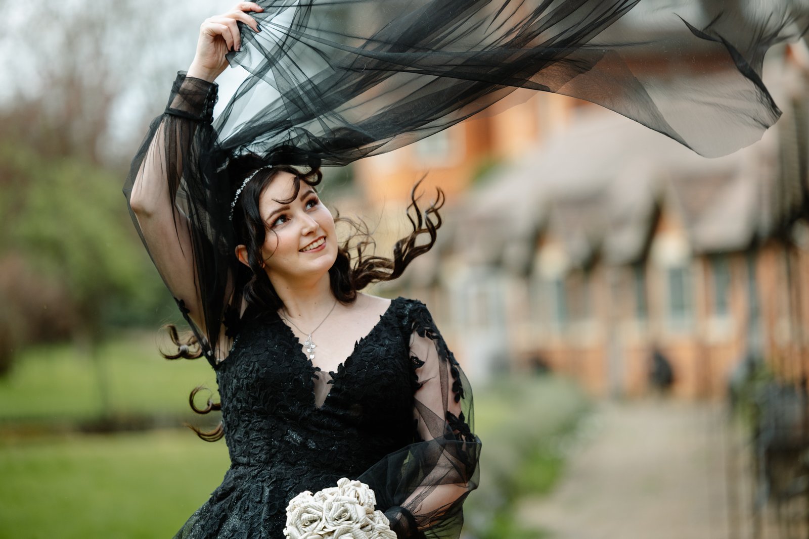 Bride in a black lace gown lifting her black veil in the breeze, thatched cottages behind her.