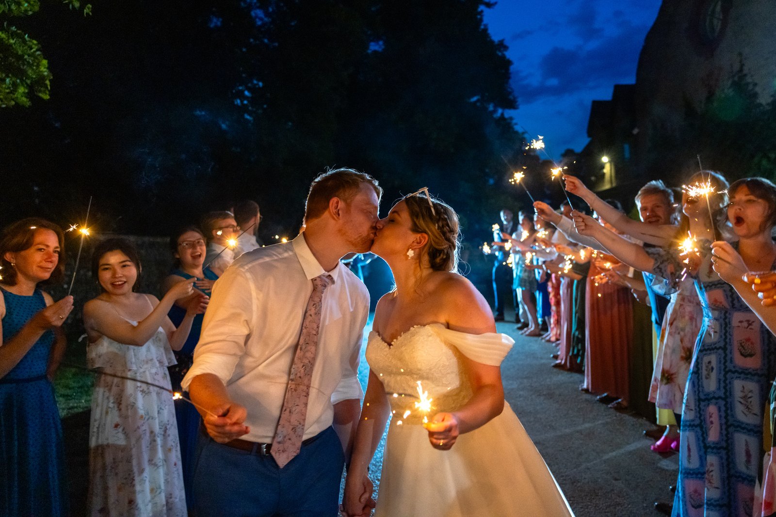 Bride and groom kissing between two lines of guests holding sparklers at night.