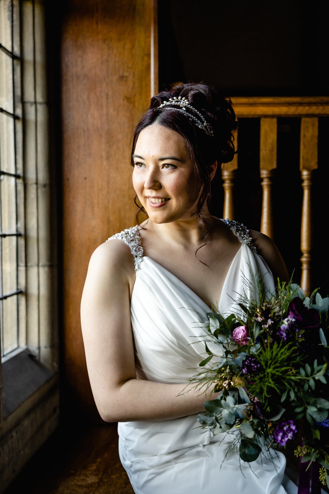 Bride pausing by a mullioned window at Fanhams Hall with a bouquet of purple and green foliage.
