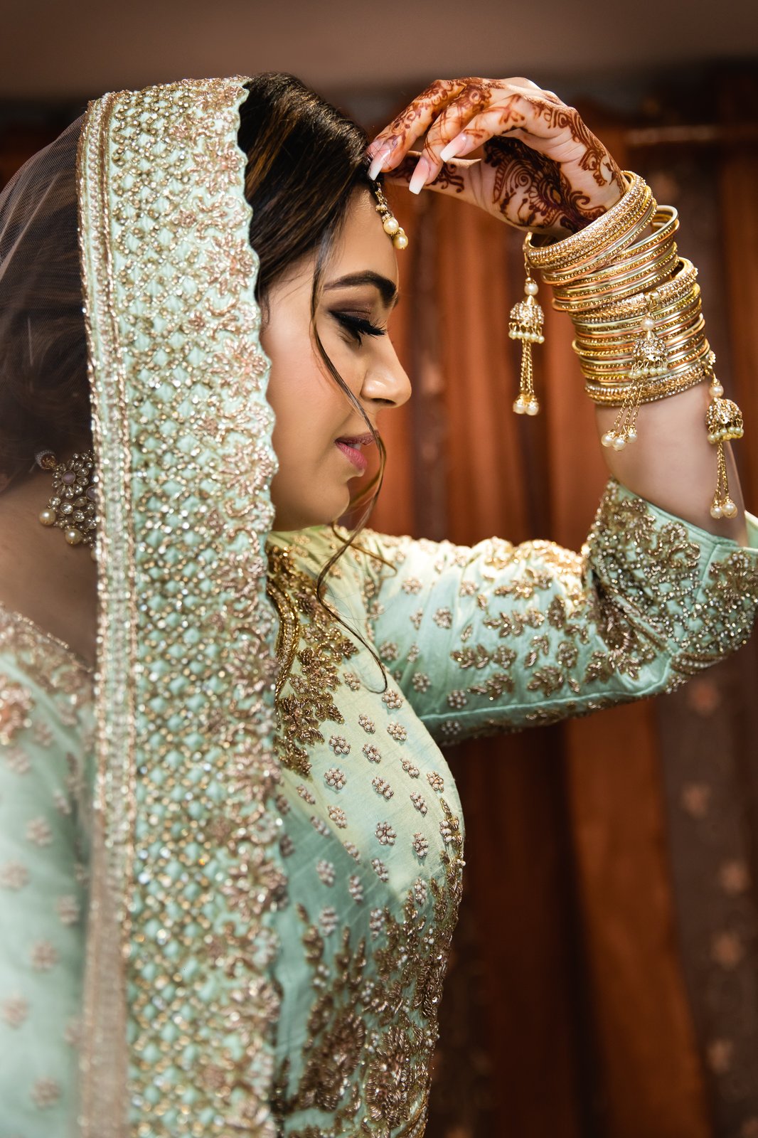 Bride in an embroidered green and gold outfit, gold bangles stacked along her arm and henna on her hands.
