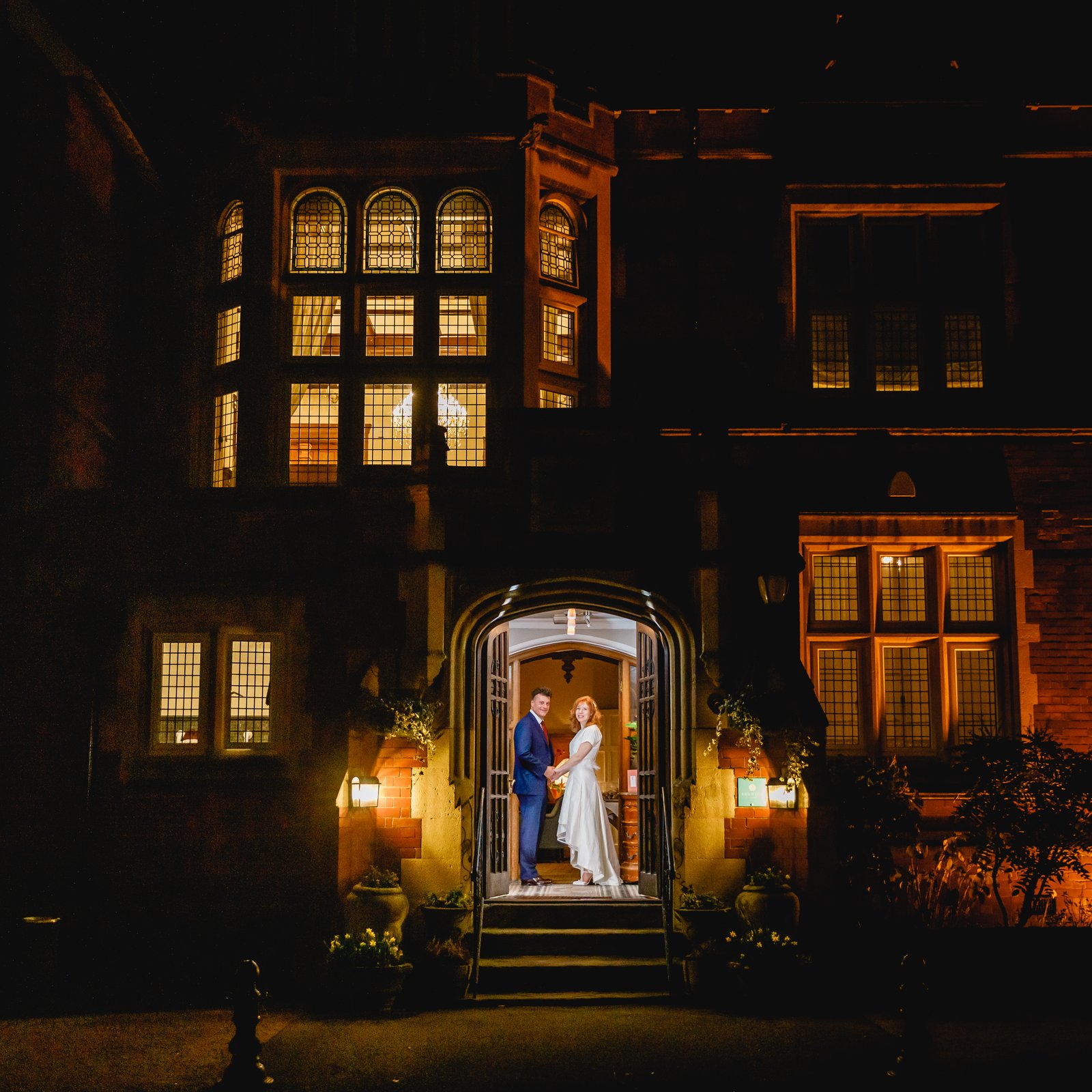 Couple standing in the lit doorway of a country house at night, its windows glowing gold.