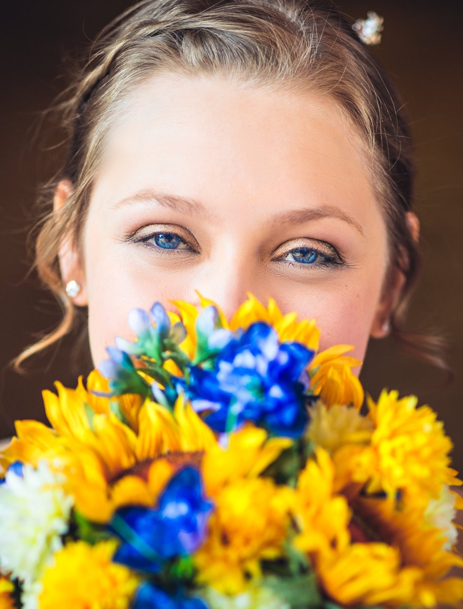 Close-up of a blue-eyed bride looking over the top of her bouquet of yellow sunflowers and blue delphiniums, her hair pinned up with a sparkling clip.