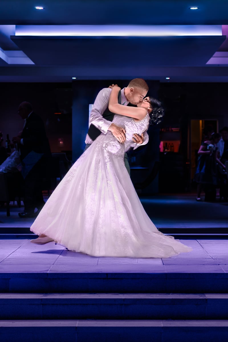 Groom dipping the bride into a kiss on a stone terrace at night, the venue lit in deep purple and blue, with guests seated at tables behind them.