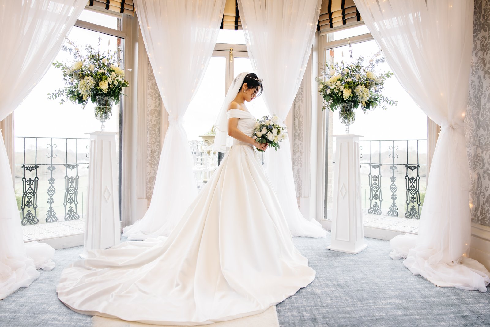Bride in an off-the-shoulder satin ball gown with a long train and veil standing between white draped curtains and tall floral arrangements by sunlit balcony windows, holding a bouquet and looking down.