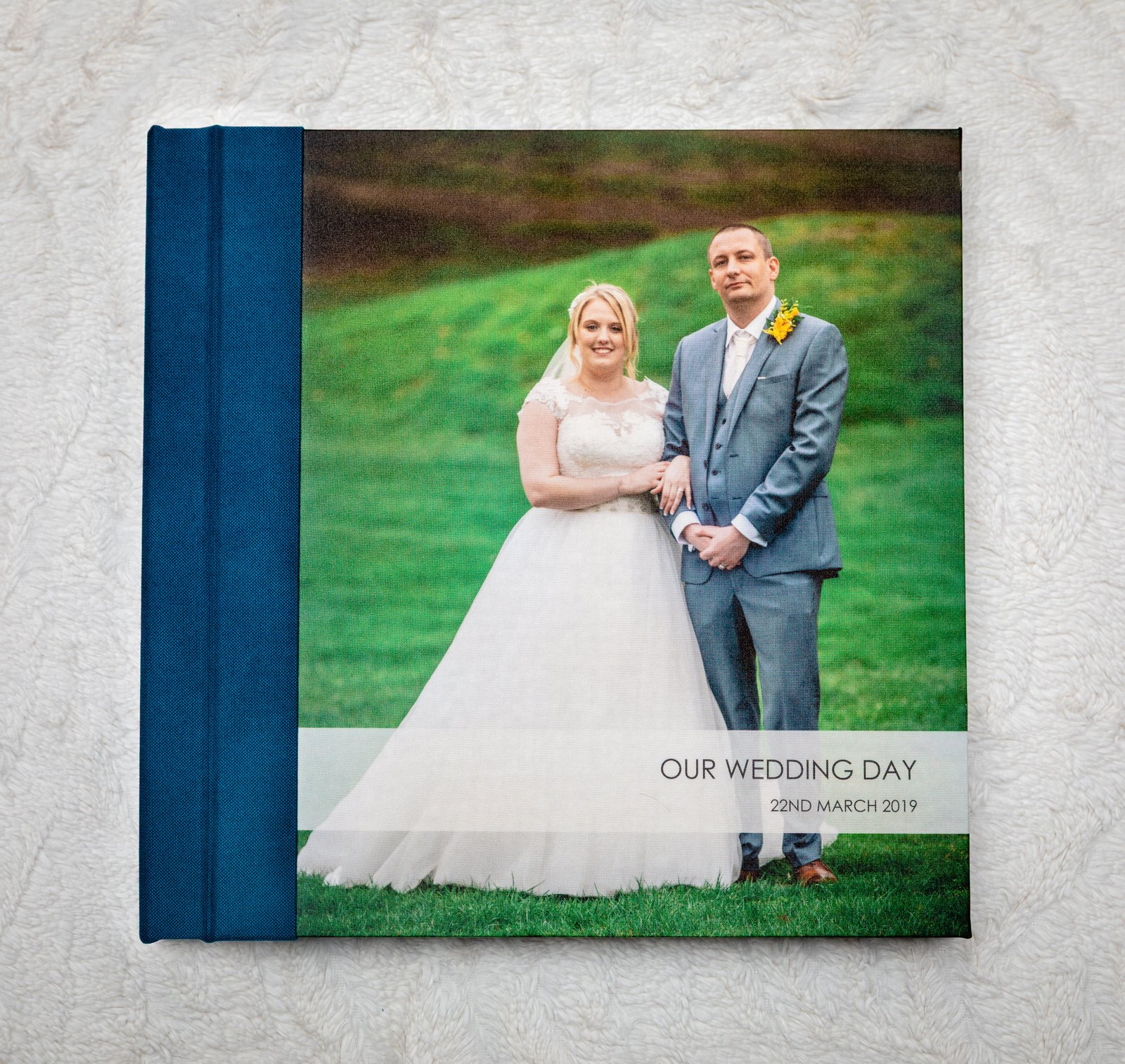 Lay-flat wedding album with a blue textile cover, the couple&rsquo;s portrait and the words Our Wedding Day printed on the front.