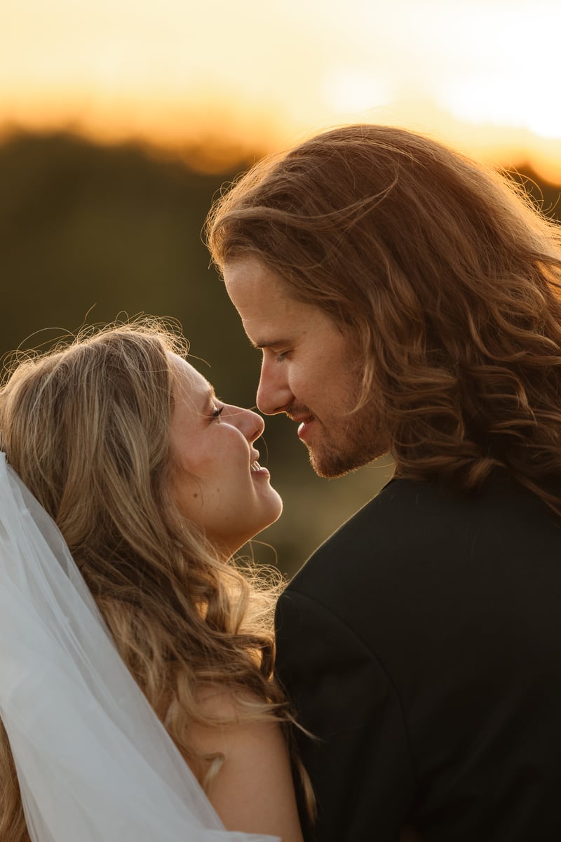 Bride and groom face to face with foreheads almost touching, smiling at each other in warm golden-hour light against a glowing sunset, the bride in a veil and the groom with long hair in a dark jacket.