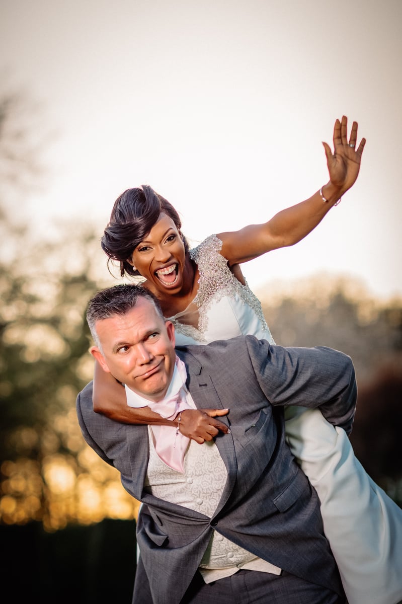 Bride laughing and waving from a piggyback on the groom&rsquo;s back, the groom pulling a deadpan face, photographed at golden hour with warm bokeh behind them.