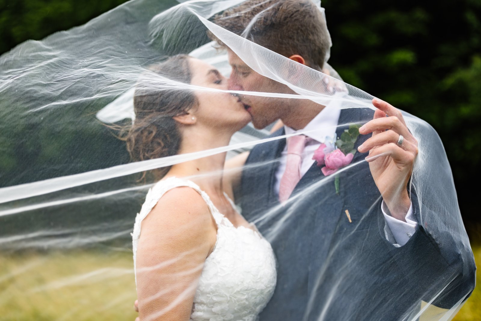 Bride and groom kissing beneath her sheer veil draped over both of them, photographed outdoors against soft green grass and trees, the groom in a grey suit with a pink tie and rose boutonnière.