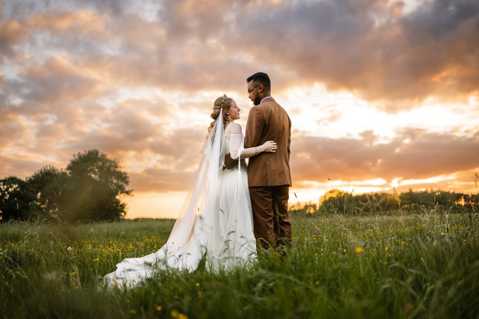 Bride in a lace gown with a long veil and groom in a brown suit standing face to face in a wildflower meadow at sunset, beneath a dramatic cloud-streaked golden sky.