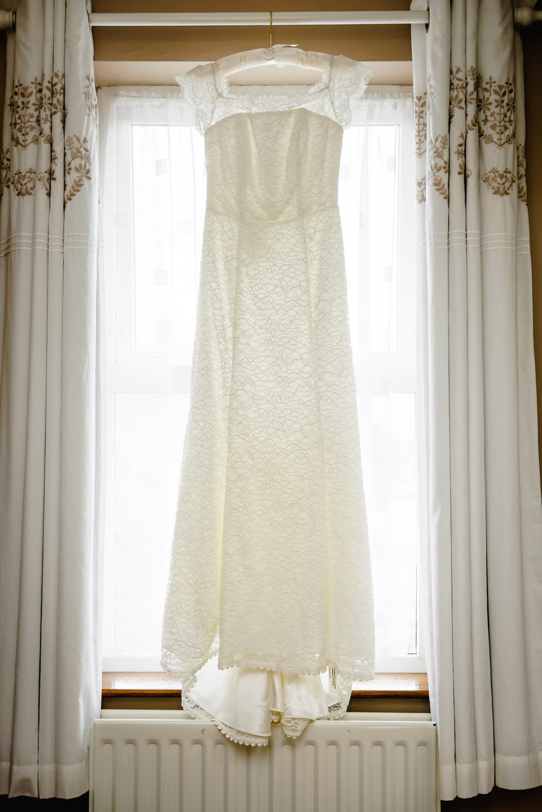 Ivory lace wedding dress hanging in a window with patterned curtains and soft morning light behind