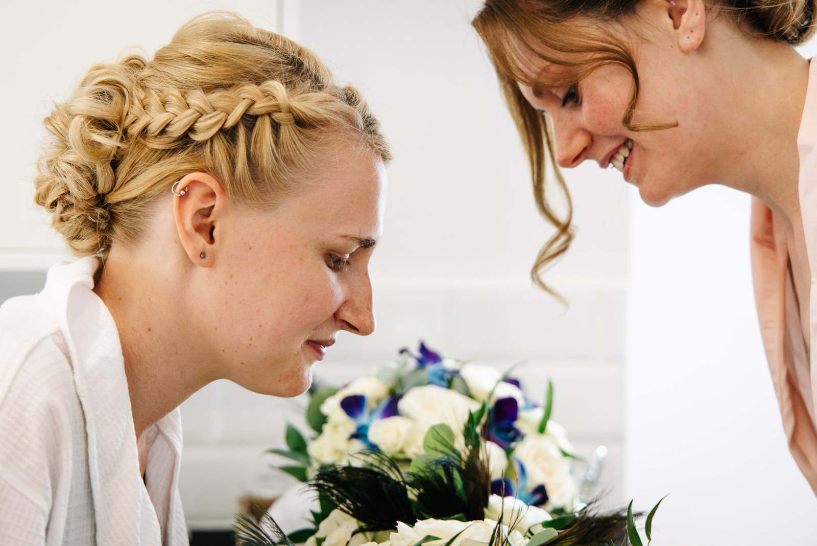 The bride leaning in to smell her white and blue bouquet beside a smiling guest