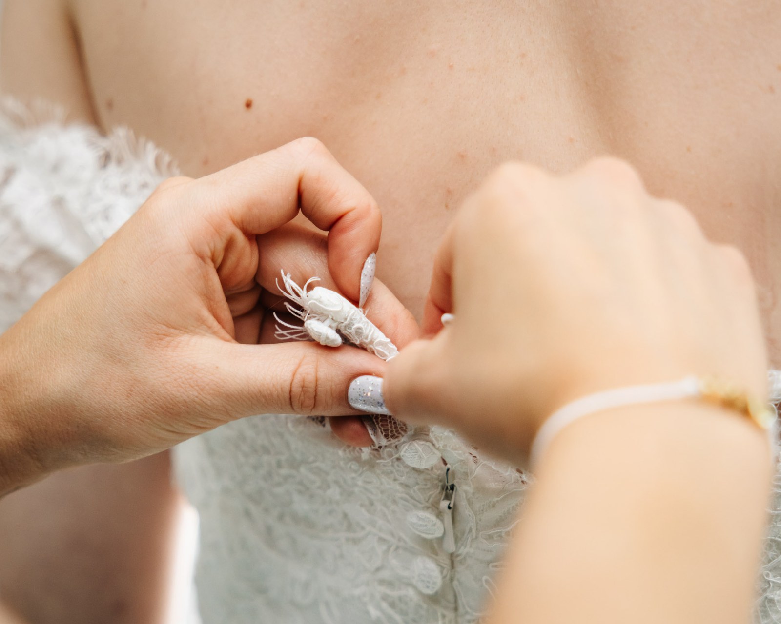 Hands fastening the back of the bride's lace gown, threading a delicate charm