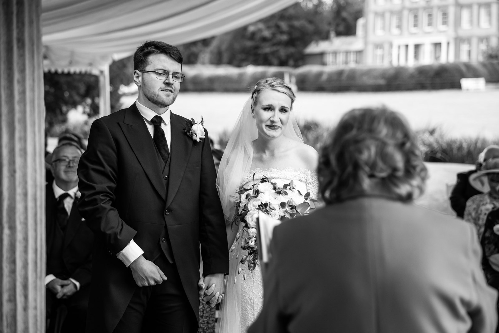 Black-and-white frame of the couple standing hand in hand at the outdoor ceremony