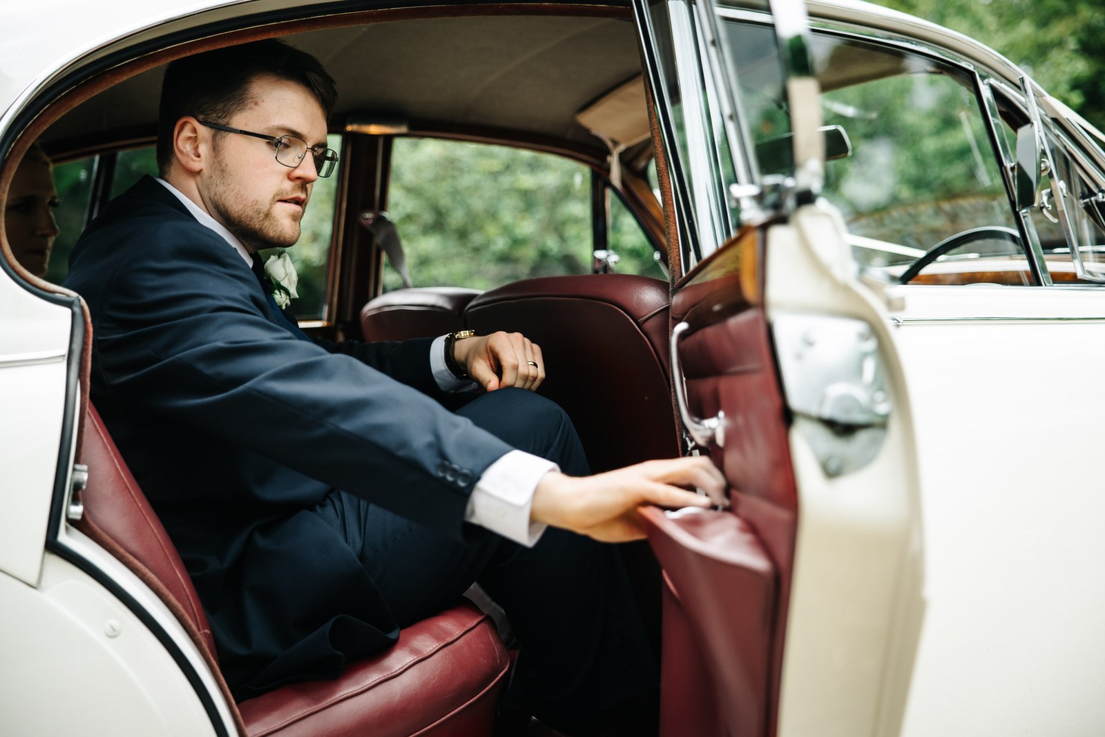 The groom seated in the red-leather interior of the vintage wedding car