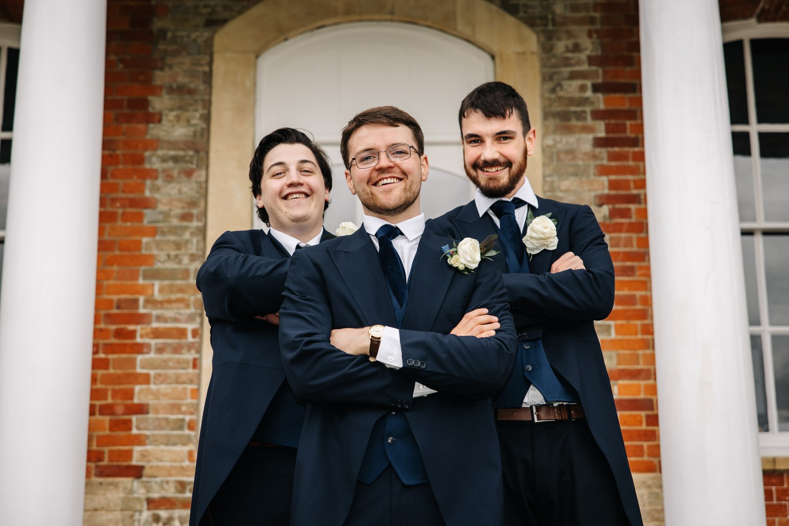 The groom with his two ushers, arms linked before the brick portico