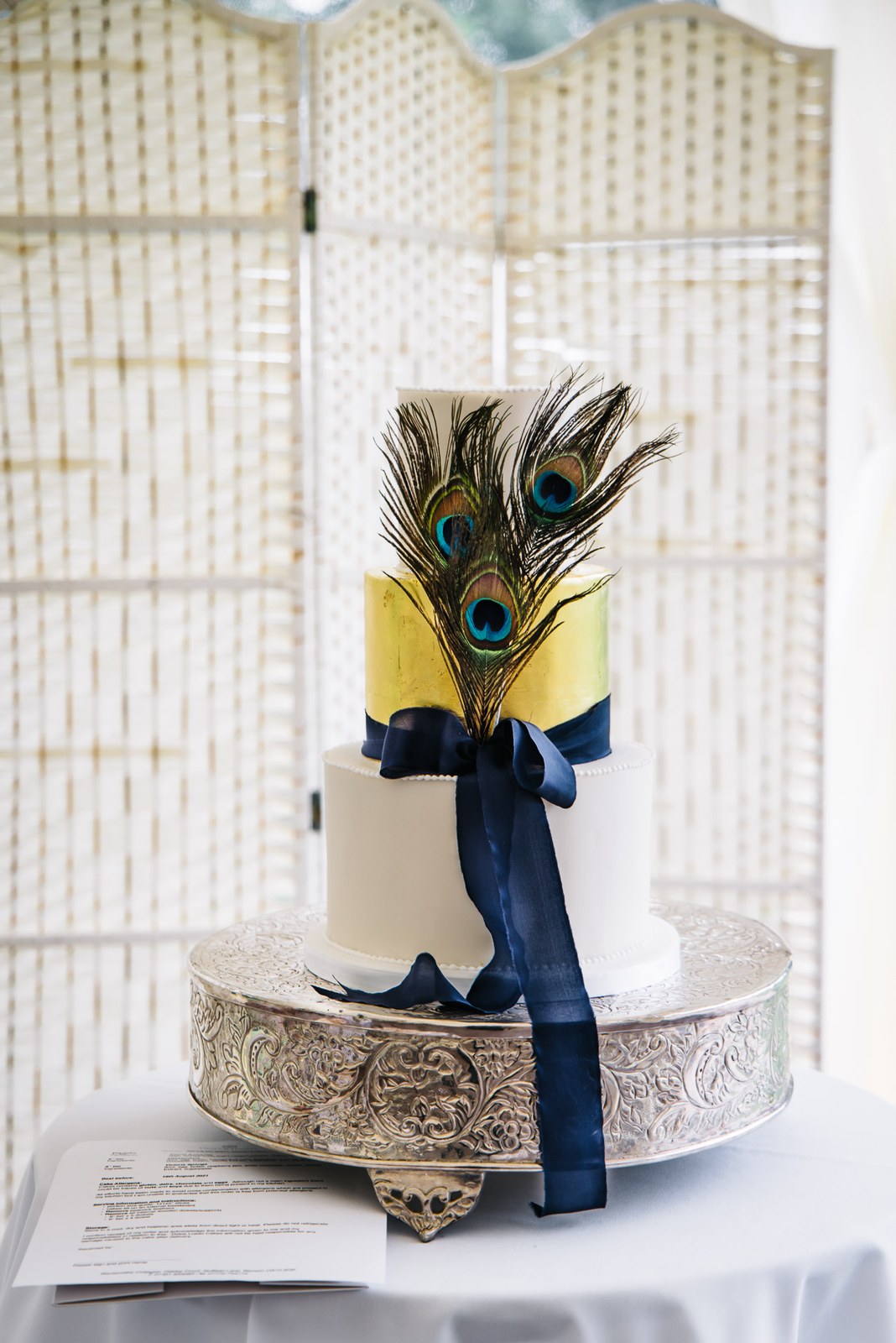 Three-tier white and gold wedding cake dressed with a navy ribbon and peacock feathers