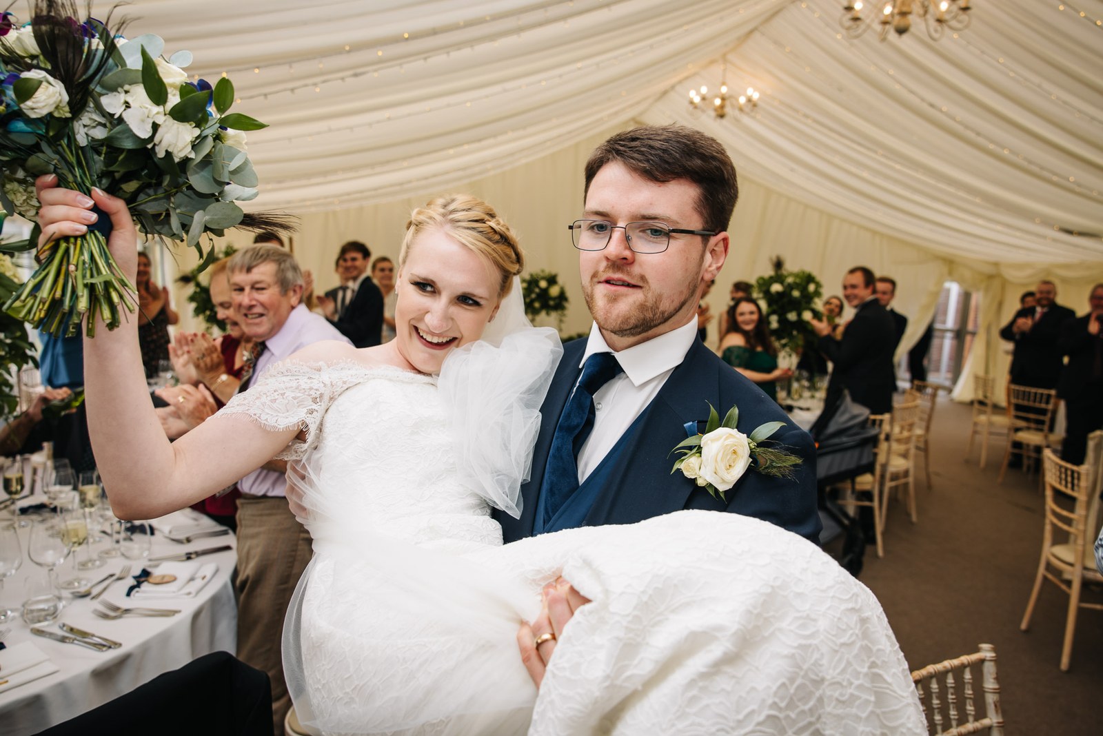 The groom carrying the bride into the marquee as guests applaud